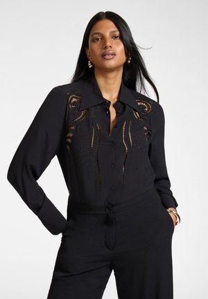 Frau mit langen schwarzen Haaren, die eine schwarze Bluse mit Ausschnittmustern und schwarze Hosen trägt, steht mit Händen in den Taschen vor einem schlichten Hintergrund.