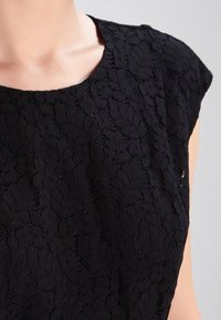 Schwarzes Spitzenoberteil mit kurzen Ärmeln, das ein florales Muster und einen runden Ausschnitt aufweist, wobei komplizierte Texturen und zarte Nähte zur Schau gestellt werden.