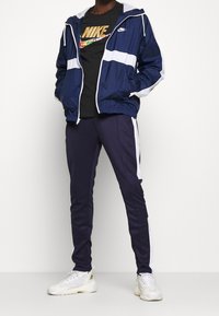 Marinblå vindjacka med vita ränder, svart T-shirt med guld Nike-logga och marinblå träningsoverallsbyxor med vita sidostripes, bärs med vita sneakers.