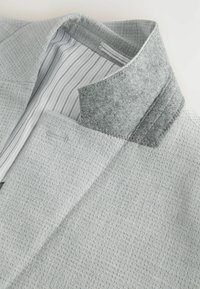 Chaqueta gris claro con una superficie tejida texturizada y un cuello de fieltro. El forro interior presenta finas rayas verticales. Costuras detalladas visibles.