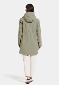 Olivgrön vattentät parka med huva, insvängd midja, långa ärmar och rak skärning, kombinerad med ljusa byxor och skor.