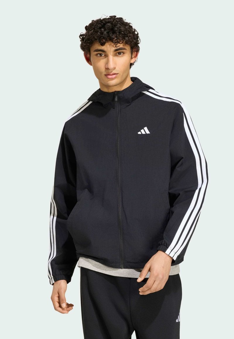 Νέος άνδρας που φοράει μαύρο μπουφάν Adidas με λευκές ρίγες στα μανίκια και γκρι πουκάμισο από κάτω, στέκεται μπροστά σε φωτεινό φόντο.