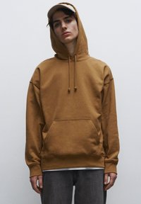 Felpa in cotone marrone con tasca a marsupio, cappuccio con coulisse e polsini a costine. Indossata sopra jeans grigi, con un cappellino chiaro.