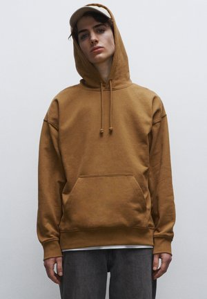 Felpa in cotone marrone con tasca a marsupio, cappuccio con coulisse e polsini a costine. Indossata sopra jeans grigi, con un cappellino chiaro.