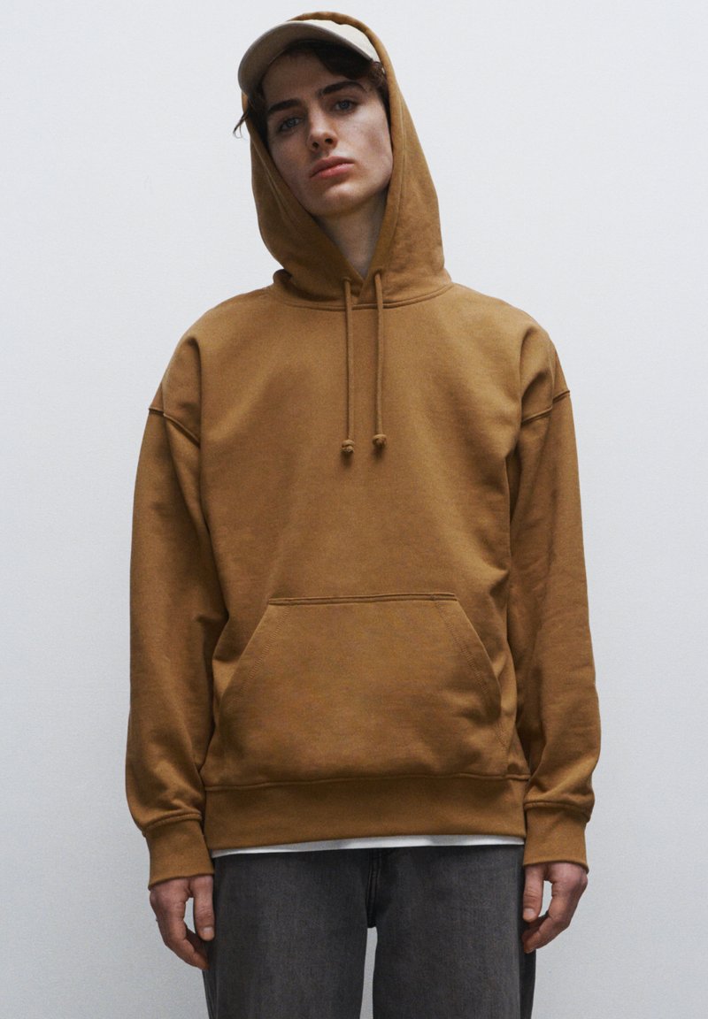 Felpa in cotone marrone con tasca a marsupio, cappuccio con coulisse e polsini a costine. Indossata sopra jeans grigi, con un cappellino chiaro.