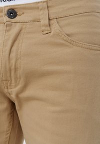 Gros plan sur un pantalon beige montrant la poche avant, la ceinture avec passants pour ceinture, et un bouton noir avec le texte "AMES" en détail.