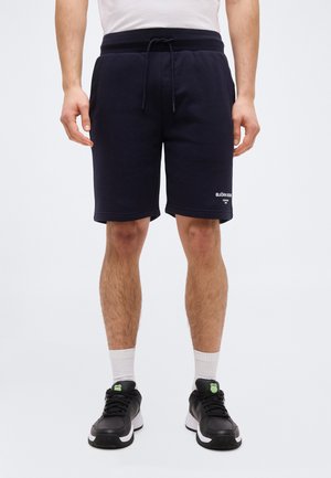 Férfi fekete zsinóros rövidnadrágban, bal lábán logóval, fehér zokniban, fekete cipőben zöld részletekkel, egyszínű háttér előtt állva.