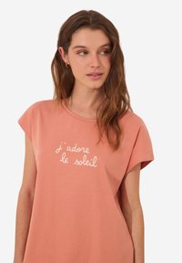 T-shirt w kolorze brzoskwiniowym o luźnym kroju, z krótkim rękawem i białym haftowanym napisem "j'adore le soleil" z przodu. Miękka bawełniana faktura.