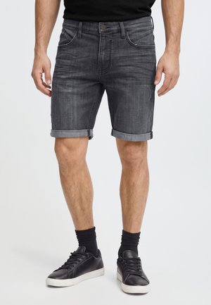 Homme portant un short en jean gris foncé retroussé, des chaussettes noires et des baskets noires sur un fond clair uni.