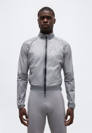 Bărbat purtând o jachetă cu fermoar și guler înalt, de culoare gri deschis, și colanți pentru ciclism căptușiți, asortați, pe un fundal simplu.