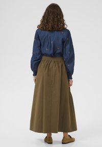 Kvinde med krøllet brunt hår iført en blå bluse med sommerfuglemønster, olivengrøn lang nederdel og beige slip-on sko, stående med ryggen til.