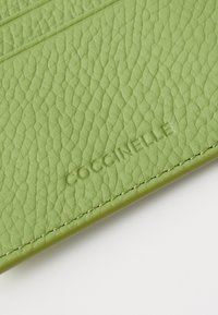 Accessorio in pelle verde texturizzata con logo "COCCINELLE" impresso, e dettagli di cucitura rifiniti lungo i bordi.