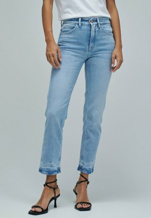 Mujer con vaqueros de talle alto de color azul claro con dobladillos remangados y sandalias negras de tacón alto con tiras, de pie frente a un fondo liso.