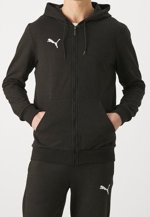 Sudadera negra con cremallera completa, bolsillos delanteros y capucha, combinada con pantalones negros, ambos con el logo blanco de Puma en el pecho y el muslo.