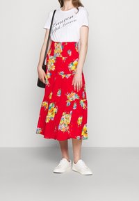 Rotes Blumenkleid in Midilänge mit gestuftem Design, kombiniert mit einem weißen T-Shirt mit Textgrafik und weißen Sneakers. Eine schwarze Schultertasche wird gehalten.
