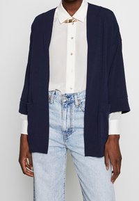 Mörkblå kofta med öppen framsida, trekvartsärmar och framfickor, lagd över en vit skjorta med knappar och ljusblå jeans.