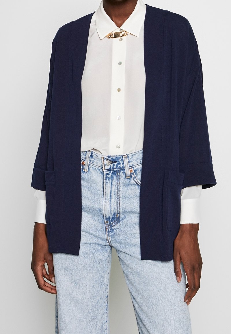 Mörkblå kofta med öppen framsida, trekvartsärmar och framfickor, lagd över en vit skjorta med knappar och ljusblå jeans.