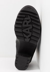 Suela de zapato de goma negra con un patrón texturizado y estriado, y un logotipo elevado que dice "KARL". Presenta una transición de talón suave para mayor estabilidad.