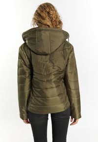 Giacca a vento verde oliva con cappuccio, caratterizzata da cuciture orizzontali e una superficie liscia e lucida. I jeans neri completano l'outfit.