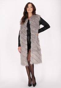 Langer grauer Kunstpelz-Westen mit einer geschichteten, wellenartigen Textur. Sie hat ein kragenloses Design und wird mit einem schwarzen Outfit und High Heels kombiniert.