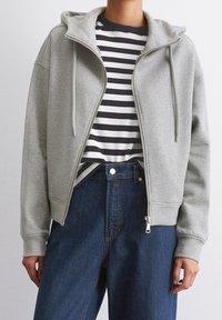 Sweat à capuche gris zippé avec une capuche spacieuse, des cordons de serrage et des poignets côtelés, superposé sur une chemise à rayures bleues et blanches, associé à un jean bleu taille haute.