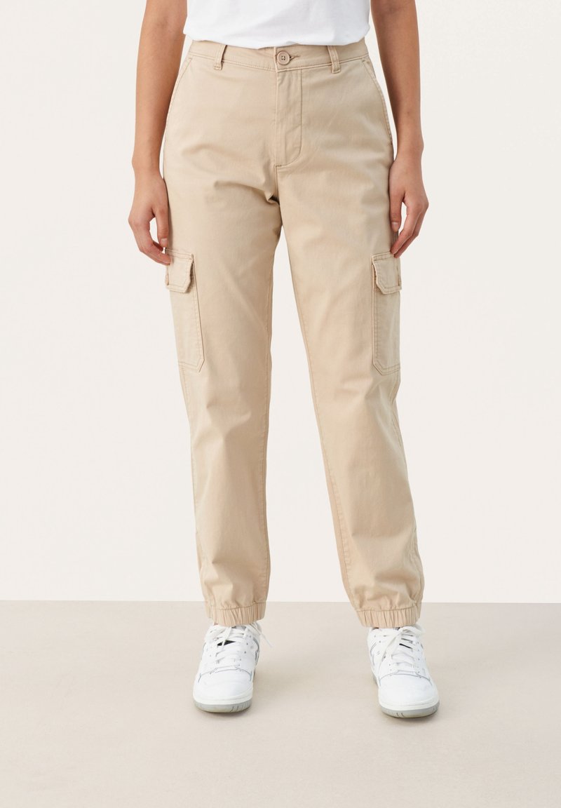 Pantalon cargo beige avec poches à rabat latérales, poignets élastiqués, porté avec des baskets blanches sur un fond neutre.