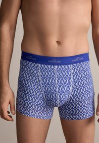 Blaue gemusterte Boxershorts mit einem weißen abstrakten Design. Der elastische Bund zeigt den Text "NEXT SIGNATURE" in einer kontrastierenden Farbe.