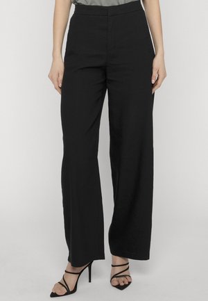 Kobieta w czarnych, szerokich spodniach z wysokim stanem, zestawionych z czarnymi, sandałami na wysokim obcasie z paskami, stojąca na jednolitym tle.