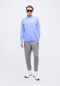 Νέος άνδρας που στέκεται φοράει μοβ μακρυμάνικη μπλούζα, γκρι αθλητικό παντελόνι, λευκά αθλητικά παπούτσια και μεγάλα κόκκινα αθλητικά γυαλιά ηλίου με απλό φόντο.