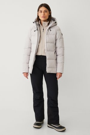 Lichtgrijze pufferjack met een capuchon, voorzien van ritszakken en een gestikt patroon, gedragen over een beige trui en zwarte broek.
