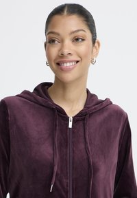 Bordowa bluza z kapturem na zamek błyskawiczny, wykonana z miękkiego materiału, z kapturem ze sznurkiem i srebrnymi elementami zamka. Charakteryzuje się luźnym krojem i brakiem widocznych wzorów.