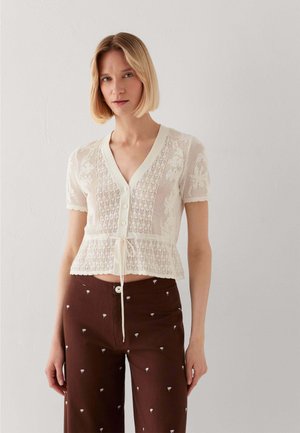 Frau mit glattem blondem Haar trägt ein weißes, gestricktes, kurzärmeliges Hemd mit Knöpfen und braune Hose mit kleinen weißen Blumenmustern.