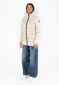 Chaqueta acolchada de un beige claro con capucha de piel sintética, cremallera negra y puños elásticos. Llevada con unos vaqueros de pierna ancha azules y zapatillas blancas.