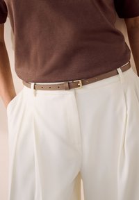 Top marrón de manga corta combinado con pantalones plisados blancos y un cinturón marrón claro con hebilla dorada. Texturas de tela suaves.