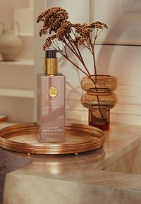 Glasflaska med rosa nyans, gulddetaljer och en pumpkork märkt "Wild Fig Hand Balm" på ett kopparfat med dekorativa blommor.