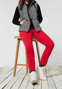 Schwarz-weißer Hahnentrittmuster-Winterweste über schwarzen Hoodie, kombiniert mit roten Hosen und weißen High-Top-Sneakern, sitzt auf einem Holzstuhl.