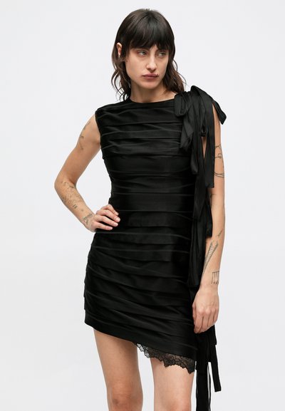 Čierne šaty s horizontálnymi skladmi, asymetrickým lemom, stuhovým detailom na jednom ramene a krajkovým akcentom na spodnej časti. Vyrobené zo saténu.