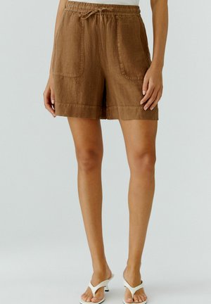 Frau trägt braune Leinen-Shorts mit Kordelzug und Fronttaschen sowie weiße Sandalen mit Absatz, steht vor einfarbigem Hintergrund.