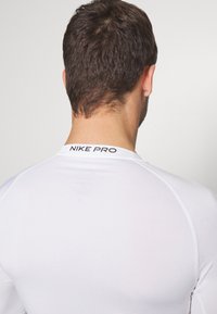 Hvid Nike Pro langærmet skjorte med rund hals, tætsiddende design og sort logo på ryggen. Glat tekstur og letvægtsmateriale.