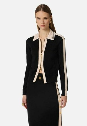 Donna che indossa una maglia nera con cerniera e strisce beige, abbinata a una gonna nera con strisce laterali beige e bottoni dorati, in piedi davanti a uno sfondo semplice.