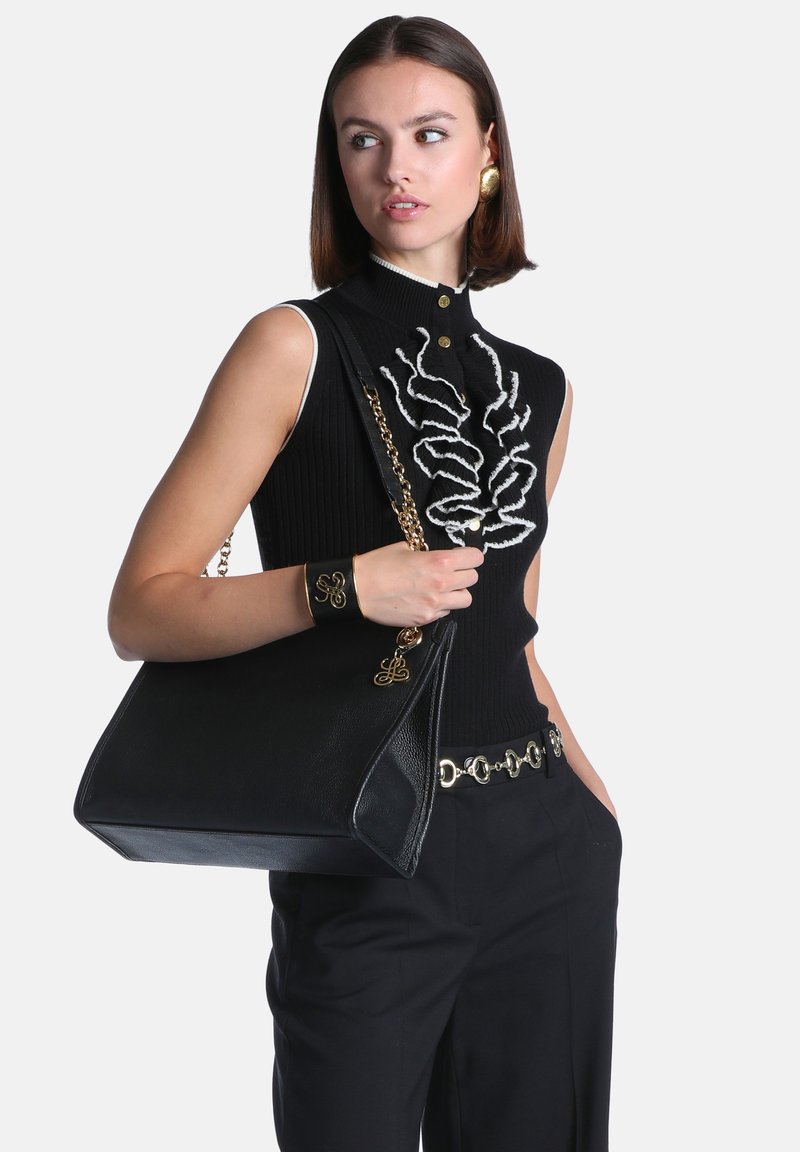Femme portant un haut noir sans manches avec des détails à volants, pantalon noir avec ceinture chaîne, tenant un grand sac à main noir avec une bandoulière chaîne dorée.