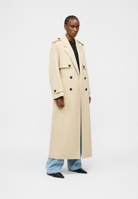 Mujer con abrigo largo beige de doble botonadura, camiseta negra, jeans anchos azul claro y zapatos negros puntiagudos, de pie frente a un fondo liso.