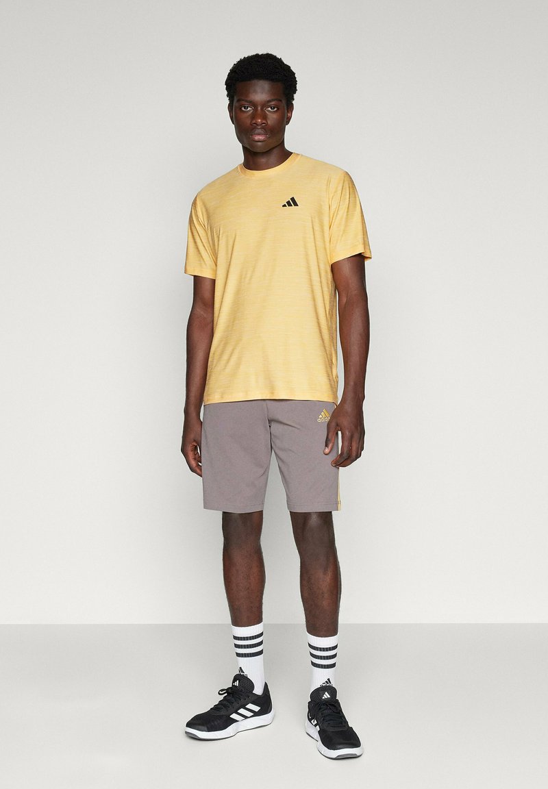 Gelb gestreiftes kurzärmliges T-Shirt mit schwarzem Logo, kombiniert mit grauen Shorts mit seitlichen Akzenten und schwarzen Sportschuhen, die mit weiß- und schwarz gestreiften Socken getragen werden.