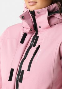 Giacca impermeabile rosa con zip nere e velcro, colletto alto con bottoni a pressione e dettaglio del logo sulla parte anteriore. Tessuto testurizzato e resistente.