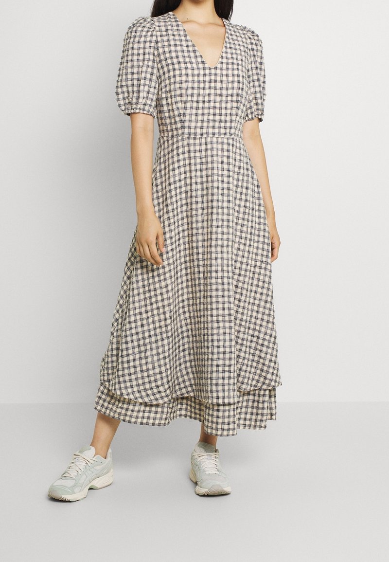 Femme portant une robe midi beige et foncée à carreaux avec des manches bouffantes et un col en V, associée à des baskets blanches, debout devant un fond uni.