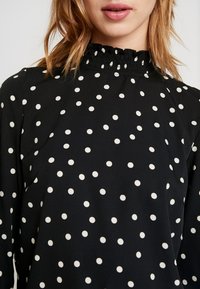 Blusa nera con pois bianchi, caratterizzata da un collo alto con volant e maniche lunghe. Tessuto morbido, con motivi distribuiti in modo uniforme.