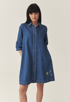 Femme portant une robe chemise en denim bleu avec des manches trois-quarts et une petite broderie florale près de l'ourlet, debout devant un fond uni.