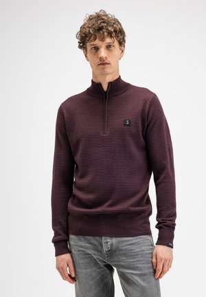 Mann mit lockigem Haar, der einen strukturierten dunkelvioletten Pullover mit Viertel-Reißverschluss und hellgraue Jeans trägt, steht vor einem schlichten weißen Hintergrund.