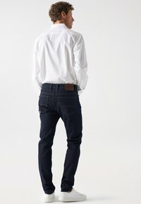 Vaqueros de mezclilla azul oscuro con un corte ajustado, que presentan dos bolsillos traseros y una etiqueta de cuero marrón en la cintura. Combinados con una camisa blanca.