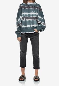 Sudadera de gran tamaño en tie-dye verde azulado con acentos grises, combinada con jeans cortos negros con desgaste y sandalias de patrón en blanco y negro.